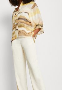 Vrouw in een losse gele en bruine marmerpatroon blouse met driekwartmouwen en wijde witte broek, met handen in de zakken.