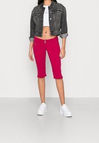 Chaqueta de mezclilla negra, diseño corto; camiseta blanca corta; pantalones capri de un vibrante color rosa con dos cierres de botón; combinados con zapatillas blancas.
