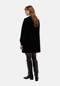 Robe en velours côtelé noir avec une coupe décontractée, de longues manches bouffantes et un col montant. Associée à des bottes noires brillantes jusqu'aux genoux et des collants noirs.