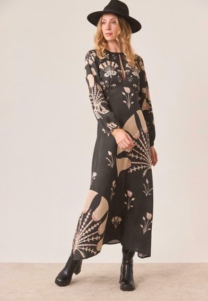 Mujer con un sombrero negro de ala ancha, vestido largo negro con estampado floral y botines negros, de pie contra un fondo liso claro.