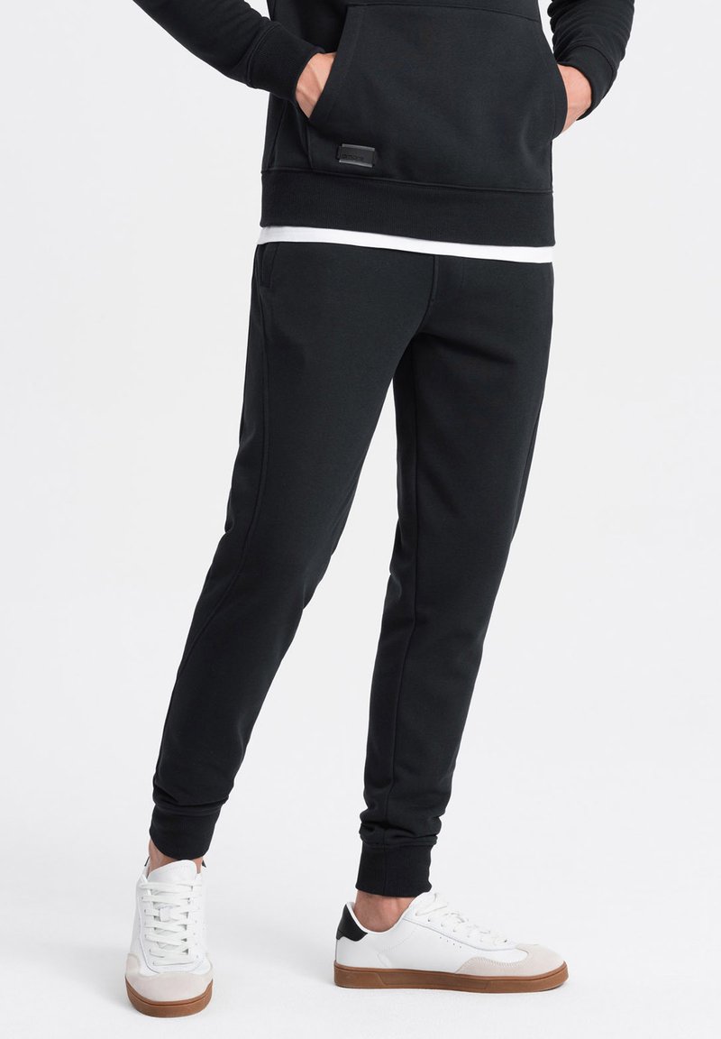 Pantalones de chándal negros con un ajuste ajustado, cinturilla elástica y bolsillos laterales, combinados con zapatillas blancas con un acento negro en el talón.