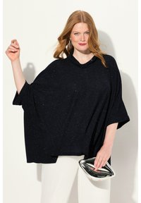 Czarna, błyszcząca, oversize'owa bluzka z dekoltem typu cowl i szerokimi rękawami, zestawiona z błyszczącą srebrną kopertówką. Modelka pozuje na jasnym tle.