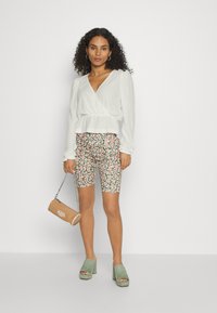 Blusa branca texturizada, calções de bicicleta com estampado floral, sandálias de salto alto verdes e uma bolsa cilíndrica bege com uma alça de corrente.