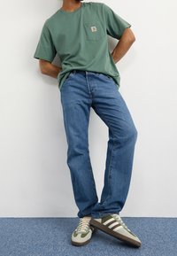 T-shirt in cotone verde con taschino sul petto, abbinato a jeans in denim blu. Le calzature presentano una combinazione di verde, beige e marrone con dettagli a righe.