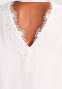Biała bluzka z dekoltem w kształcie litery V, ozdobiona misternymi koronkowymi detalami wokół dekoltu, o miękkiej fakturze materiału oraz z pojedynczym zapięciem na guzik z tyłu.