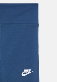 Mėlynos sportinės tamprės iš lygaus, tamprios medžiagos. Yra aukštas juosmuo ir baltas „Nike“ logotipas ant kairės pusės.