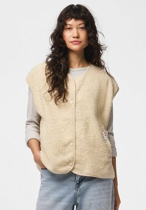 Kvinde med langt, bølget hår iført en beige fleecervest med knapper over en lysegrå langærmet skjorte og lyseblå jeans, stående med den ene hånd i lommen.