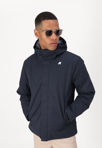 Chaqueta de invierno azul marino con capucha, cremallera frontal y logotipo en el lado izquierdo del pecho; cuenta con bolsillos frontales y textura suave.