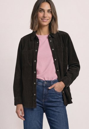 Kvinde med skulderlangt brunt hår iført en lyserød skjorte, mørkebrun kordfløjlsjakke med knapper og blå jeans med en hånd i lommen.