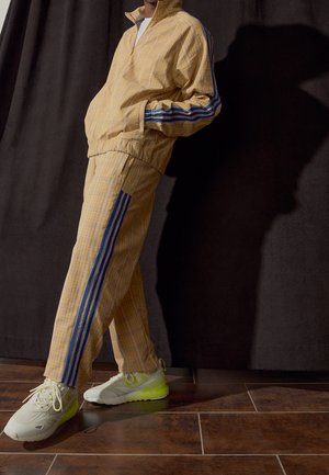 Person trägt beige Trainingsanzug mit blauen Streifen an Ärmeln und Hosen, weiße und neongrüne Sneakers, steht auf braun gefliestem Boden.