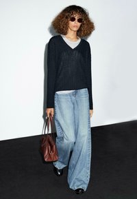 Donna con capelli ricci, che indossa occhiali da sole, un maglione nero a maglia, jeans larghi chiari e scarpe nere, mentre porta una grande borsa di pelle marrone e cammina all'interno.