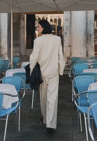 Beige tailored suit with a relaxed fit, paired with a black jacket, walking on stone pavement near blue chairs and tables.
