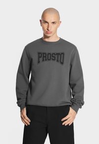 Szary sweter z okrągłym dekoltem i długimi rękawami, z czarnym, wyrazistym napisem "PROSTO" z przodu. Prosty design i materiał bawełniany.