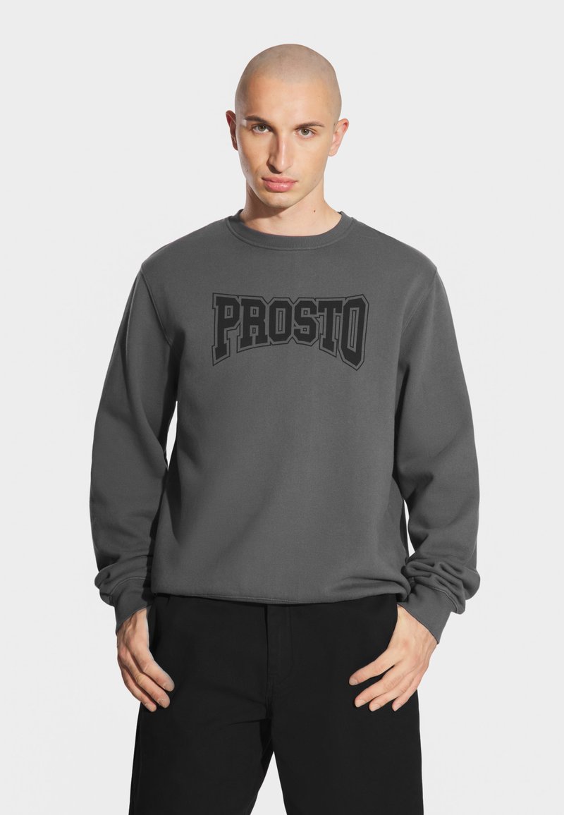 Szary sweter z okrągłym dekoltem i długimi rękawami, z czarnym, wyrazistym napisem "PROSTO" z przodu. Prosty design i materiał bawełniany.