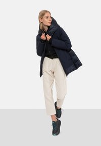 Marineblauwe donsjas met een grote capuchon, gecombineerd met een zwart shirt, crèmekleurige pantalon en zwarte sneakers met turkooizen veters en accenten.