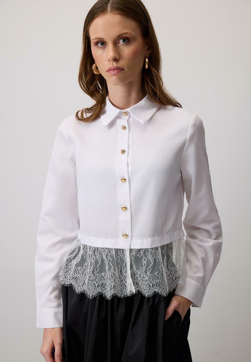 Cămașă albă cu guler, prezentând butoane aurii și un overlay din dantelă la partea de jos, cu un model floral, asortată cu o fustă neagră.
