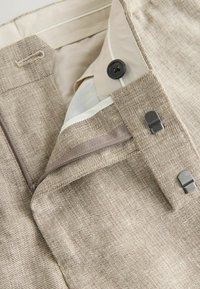 Gros plan sur un pantalon en tissu beige texturé avec un bouton noir, des crochets métalliques et une doublure beige à l'intérieur de la ceinture.
