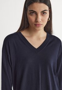 Marinblå V-ringad tröja, långa ärmar, mjuk textur, ribbad krage. Modellen bär guldfärgade hoopörhängen, har håret nedsläppt och minimal sminkning.