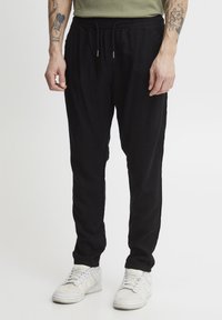 Pantalons de jogging noirs en tissu léger, dotés d'une taille élastique avec cordon de serrage, de poches latérales et de jambes fuselées. Baskets blanches.