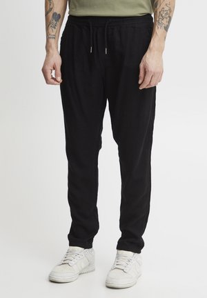 Zwarte joggingsbroek van een lichtgewicht stof, met een elastische tailleband met trekkoord, zijzakken en taps toelopende pijpen. Witte sneakers.