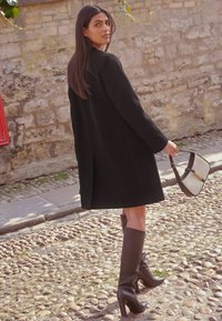 Cappotto di lana nero con taglio dritto, stivali al ginocchio in pelle marrone con tacco e borsa strutturata color crema con dettagli marroni.