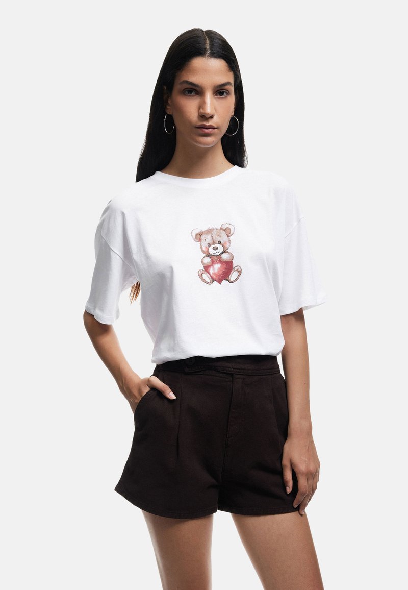 Witte katoenen T-shirt met een cartoonbeer die een rood hart vasthoudt, korte mouwen, ontspannen pasvorm, gecombineerd met donkerbruine high-waisted shorts.