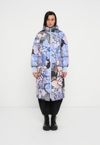 Manteau matelassé à fleurs avec capuche, présentant des couleurs bleue, rose et noire, une fermeture éclair et des poignets côtelés. Pantalon noir et bottes noires épaisses.