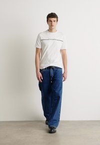 Hvid kortærmet t-shirt med grøn grafisk tekst, parret med løstsiddende blå denim jeans. Båret med sorte sko, stående mod en lys baggrund.