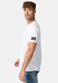 Camiseta de algodón blanca con mangas cortas, abertura lateral y etiqueta negra en la manga izquierda con el texto "//1993". Corte clásico. Jeans de mezclilla.