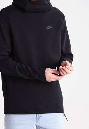 Person trägt einen schwarzen Nike-Hoodie, Hände justieren die Ärmel, hellblaue Jeans sichtbar, auf schlichtem hellem Hintergrund.