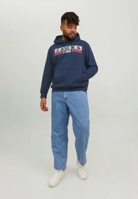 Tmavě modrá mikina s kapucí s detailem loga na přední straně, klokáním kapsou a stahovací šňůrkou na kapuci, doplněná světlými modrými džíny a bílými teniskami.