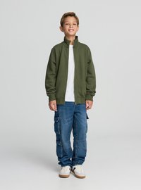 Giacca verde oliva con cerniera e collo alto, indossata sopra una maglietta bianca; abbinata a pantaloni cargo blu e sneakers bianche.