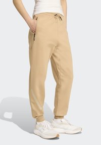 Béžové joggers vyrobené z mäkkého materiálu s elastickým pásom, sťahovacou šnúrkou, bočnými vreckami a reflexnými detailmi na nohách.