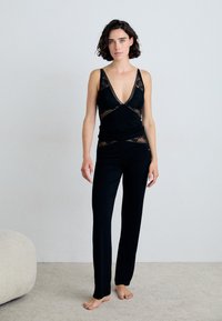 Musta rentoutumisasu, jossa on syvä V-pääntie ja pitsikoristelu sekä pitkät housut. Pehmeä kangas, jolla on sileä tekstuuri.