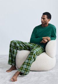 Πράσινο μακρύ μανίκι πουκάμισο σε συνδυασμό με καρό πιζάμα σε πράσινο και κίτρινο χρώμα. Καθισμένο σε μια στρογγυλή, υφή κρέμα καρέκλα πάνω σε γκρι χαλί.