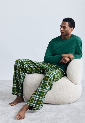Chemise à manches longues verte associée à un pantalon de pyjama à carreaux vert et jaune. Assis sur une chaise crémeuse arrondie et texturée sur un tapis gris.