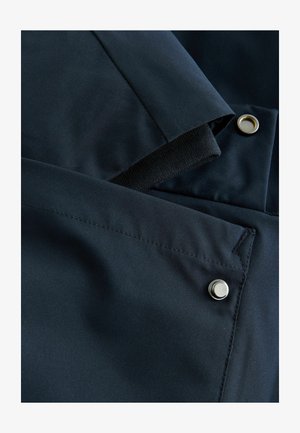 Chaqueta azul marino con una textura suave y lisa, adornada con puños elásticos negros en contraste y botones a presión plateados.