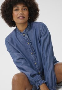 Een denim overhemd met een ruches kraag, knoopsluiting aan de voorkant en lange mouwen met elastische manchetten. De kleur is een middelblauwe denim.