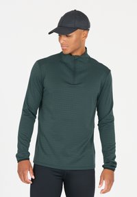 Donkergroene sporttop met lange mouwen en een kwart-rits kraag. Bevat textuur in horizontale lijnen en heeft een nauwsluitende pasvorm. Dragen met zwarte broek en een pet.