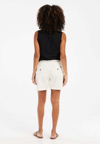 Zwarte mouwloze top, beige shorts met achterzakken, platte sandalen, krullend haar, gezien van achteren tegen een witte achtergrond.