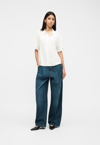 Γυναίκα με μακριά σκούρα μαλλιά που στέκεται με τα χέρια στις τσέπες, φοράει άνετο λευκό κοντομάνικο πουκάμισο, φαρδιά μπλε τζιν και μαύρα παπούτσια.