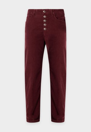 Femme aux longs cheveux blancs portant un blazer noir avec bordure en dentelle, un haut blanc, un pantalon bordeaux avec des boutons devant et des talons noirs, debout sur un fond blanc.