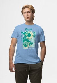 Camiseta de algodón azul claro con un estampado gráfico de girasoles, texto "Vincent" y "Amsterdam VAN GOGH" con acentos verdes.