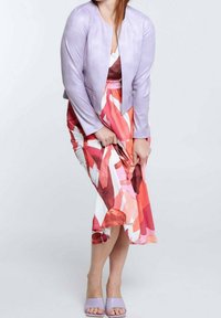 Veste en cuir lavande claire avec poches zippées, portée par-dessus une robe à motifs floraux colorés dans des tons de rose et corail.