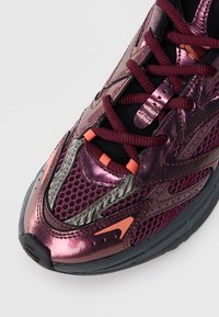 Baskets de sport violettes avec une finition métallique brillante, des panneaux en maille, des accents oranges et des lacets noirs. Elles sont dotées d'une semelle noire rembourrée.