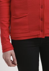 Röd stickad cardigan med knäppning fram, långa ärmar och en liten ficka. Texturerad tyg med ribbade ärmslut och nederkant. Svarta byxor synliga.