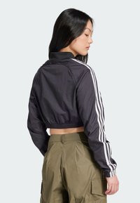 Orezaná čierna bunda vyrobená z ľahkého materiálu, s bielymi pruhmi na rukávoch a rebrovaným lemom; doplnená o olivové cargo nohavice.