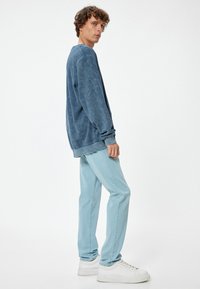 Sudadera azul de gran tamaño en tejido suave, combinada con jeans de un azul claro. El modelo se presenta de lado, mostrando el corte holgado y las mangas ajustadas.