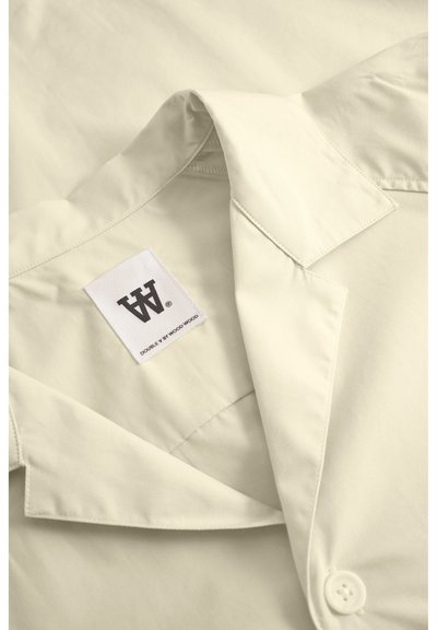 Chemise couleur crème avec un col classique, un devant boutonné et une étiquette affichant "DOUBLE V BY WOOD WOOD." Texture de tissu lisse, design minimaliste.