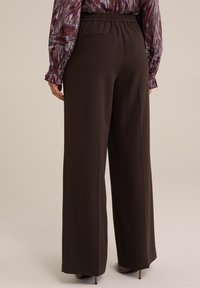 Woman wearing dark brown wide-leg trousers and a long-sleeve patterned blouse, standing against a plain beige background.